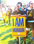 A young Black man stands in a crowd on a lawn holding a sign that says "I AM Human"