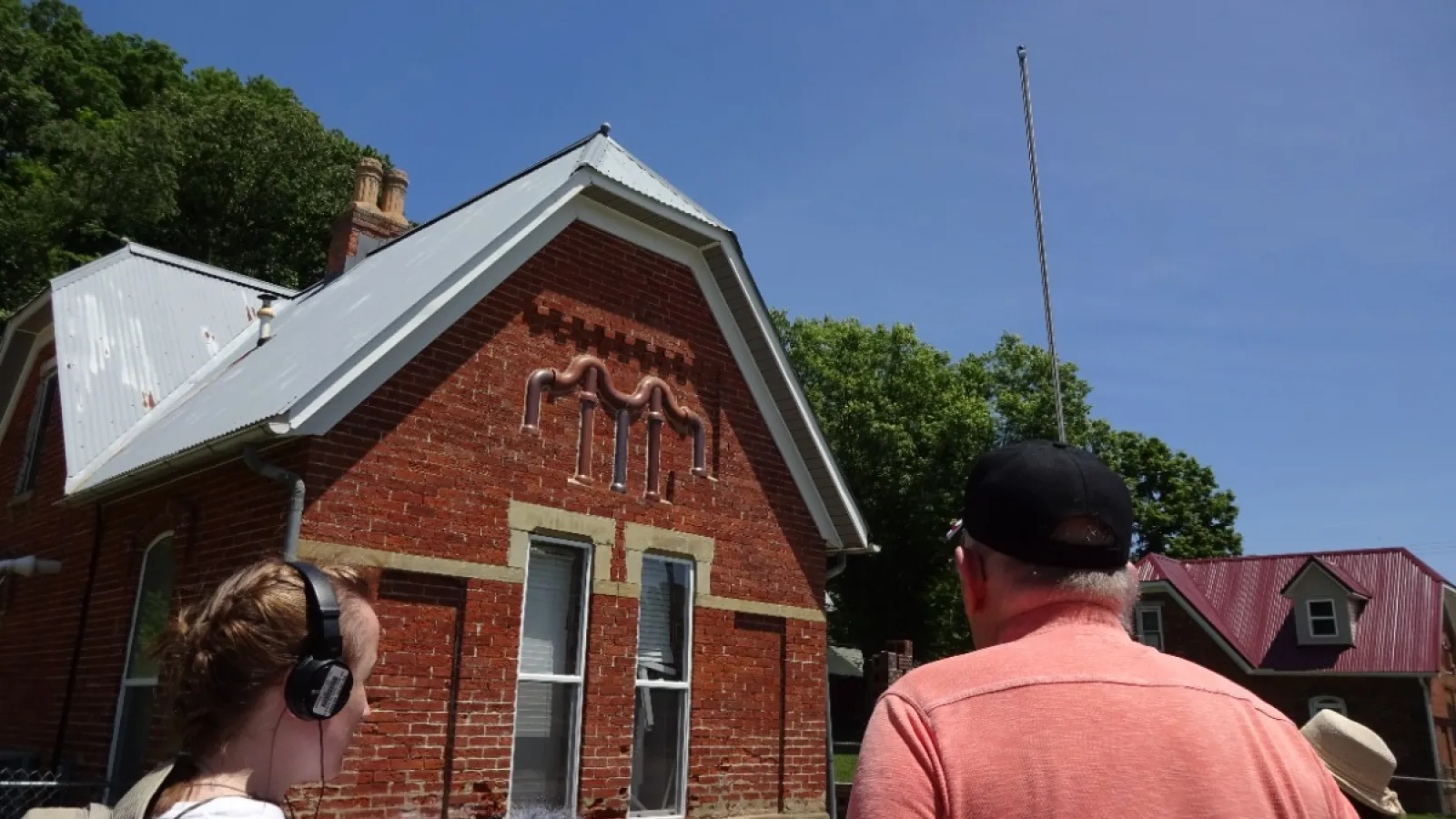 Emily Kovacic, Larry Horn, and a woman with a hat gathered in front of a brick house with clay pipe decorating the front of it.