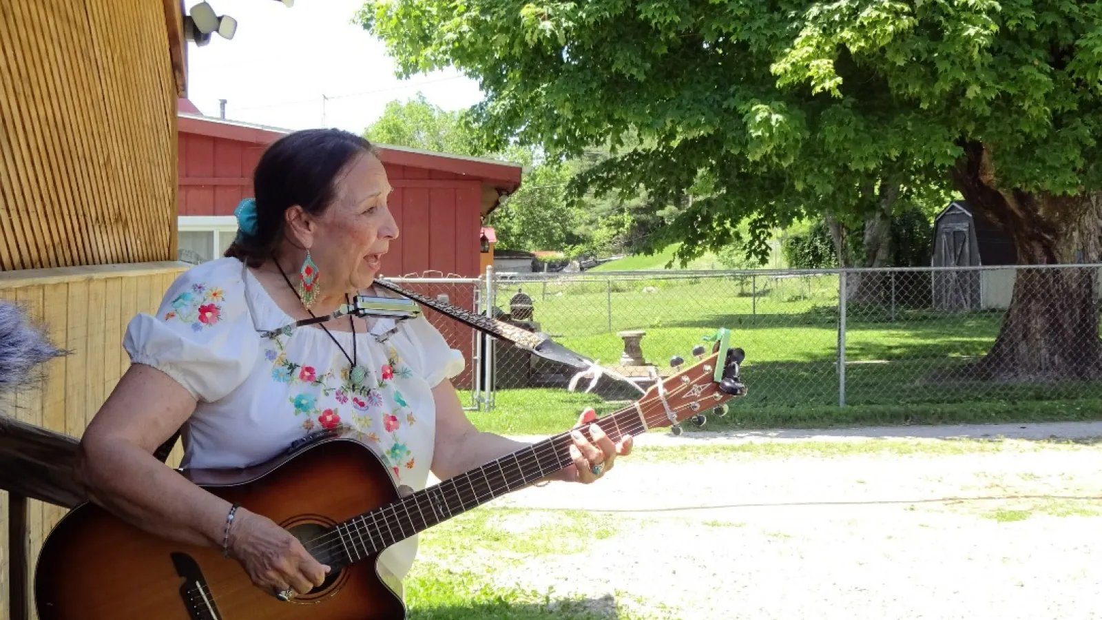 Nancy Devol Rose performing, playing her acoustic guitar with a harmonica around her head.