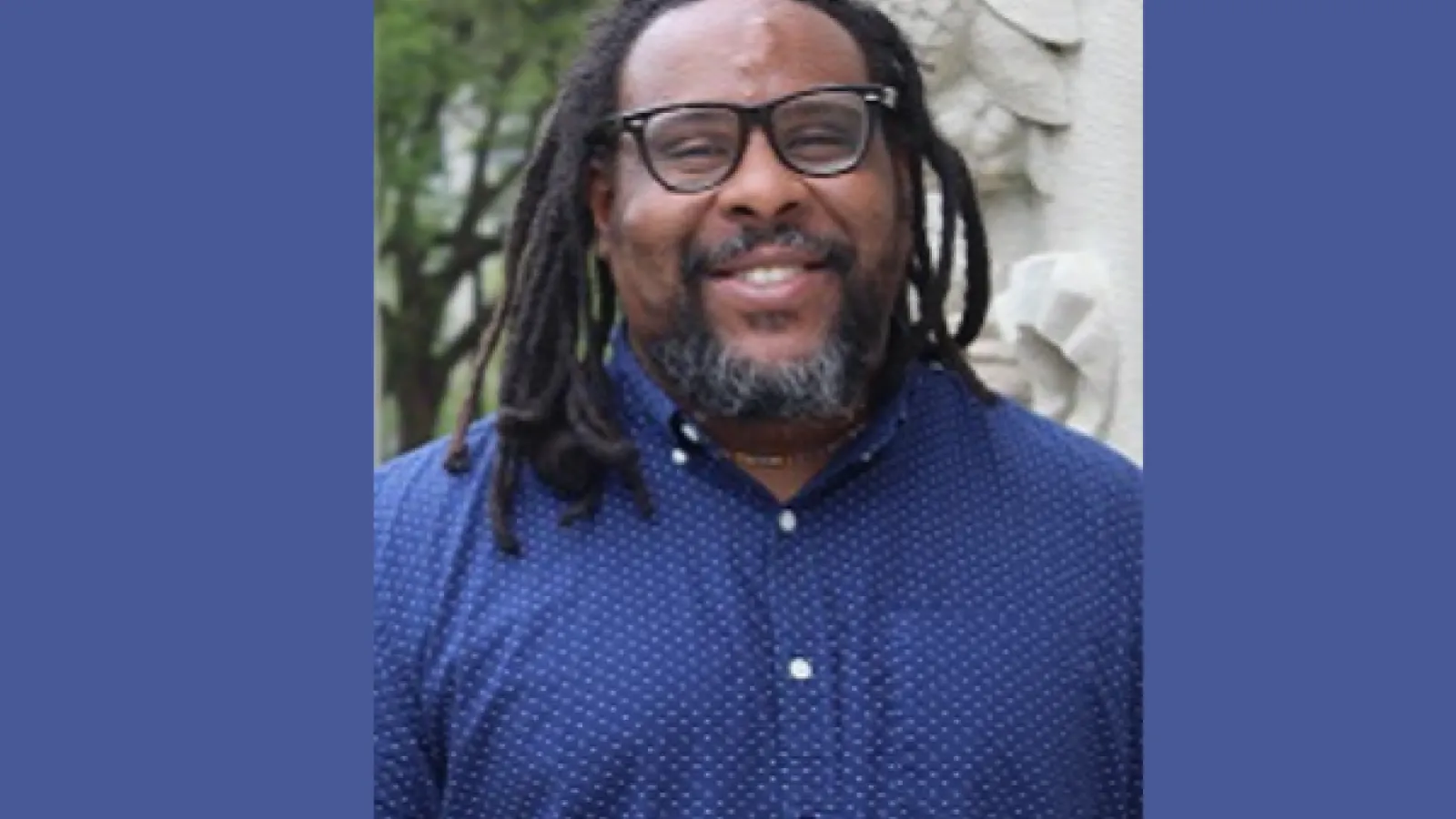 Portrait of David Todd Lawrence. he is wearing a purple button down shirt and glasses. he has dreadlocks and a beard.