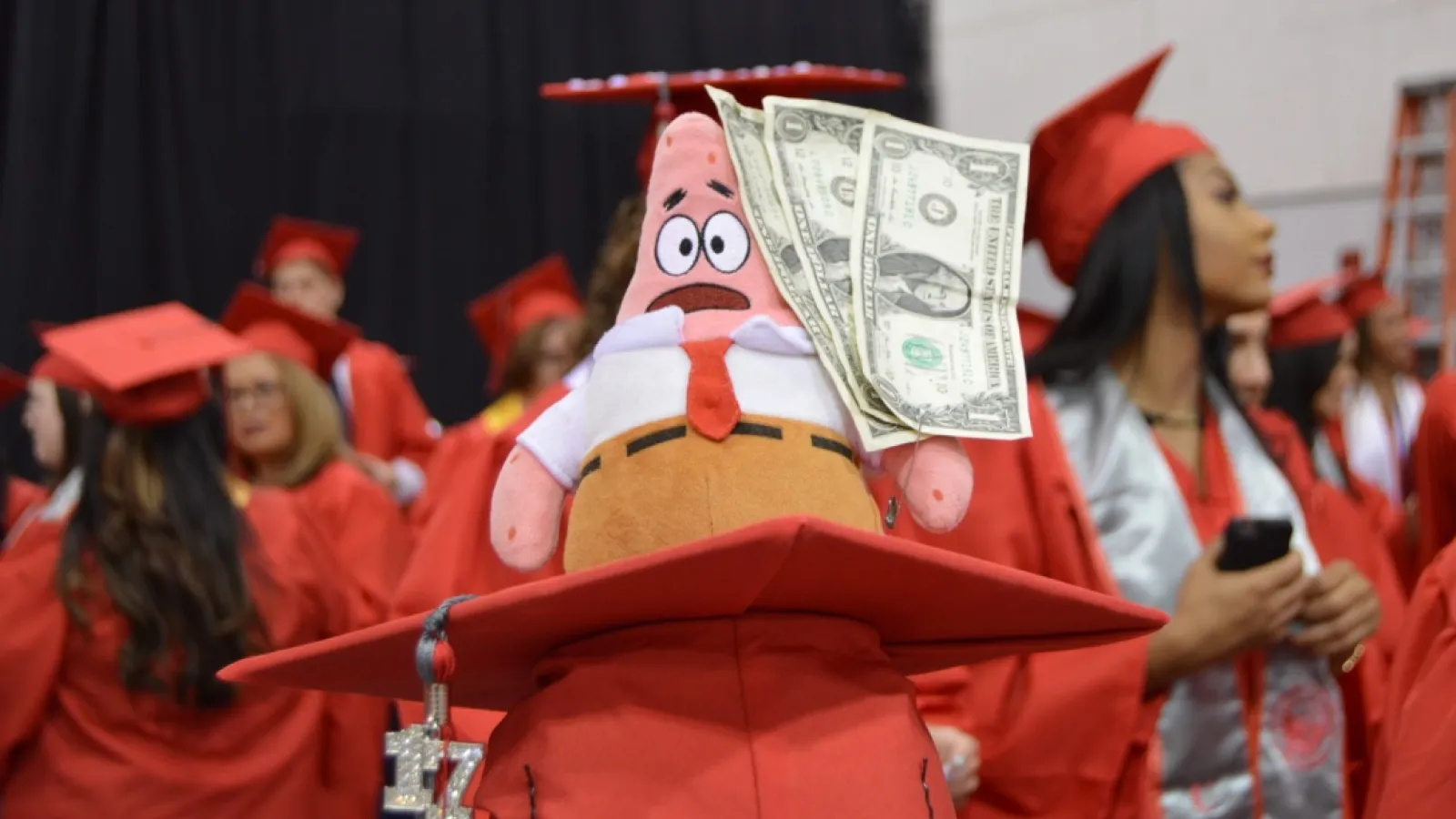 A graduating student wearing a mortarboard cap with Patrick Star dressed in collared shirt with tie and holding three one dollar bills