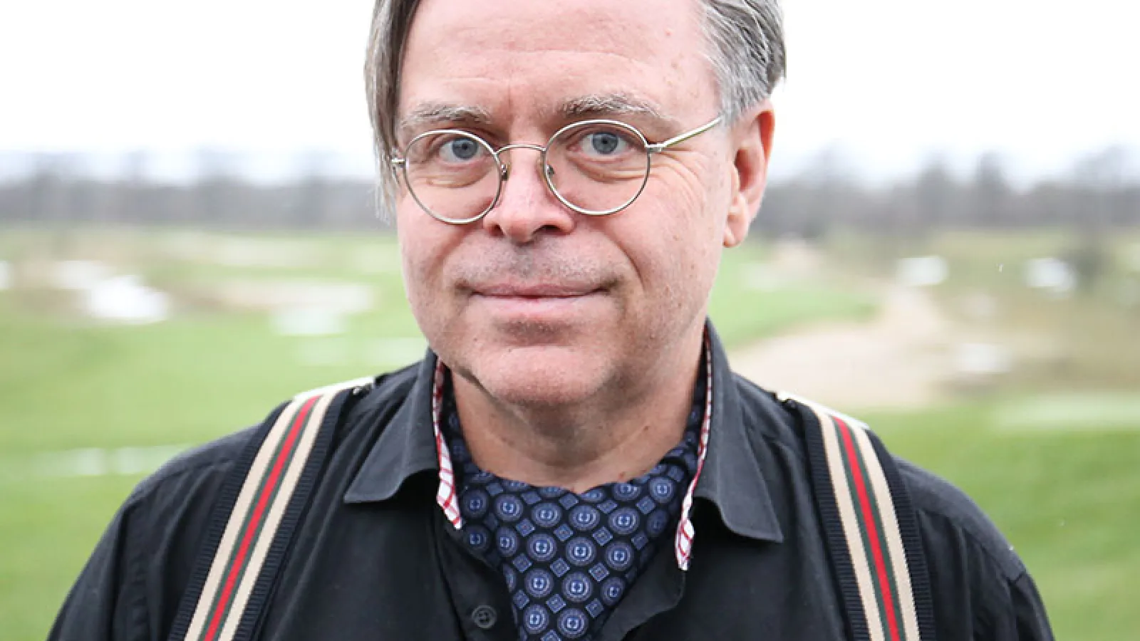 Older man with gray hair, round glasses, wearing a blue button up shirt with striped suspenders