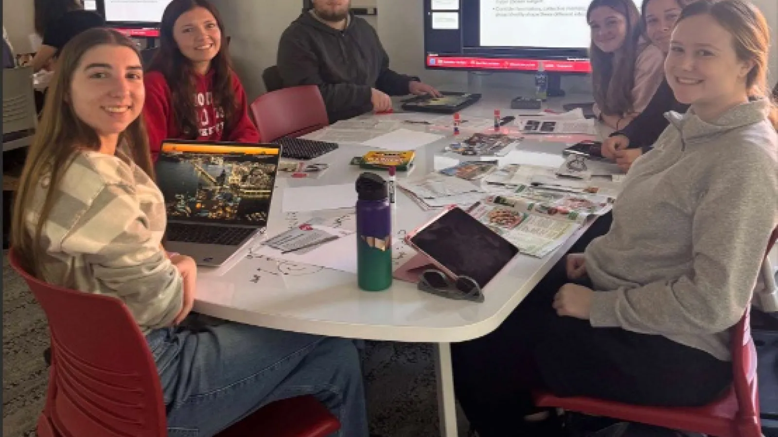 A group of students sitting around a table working on a collage. Students are looking at the photographer. There is a screen in the background with information regarding the task.