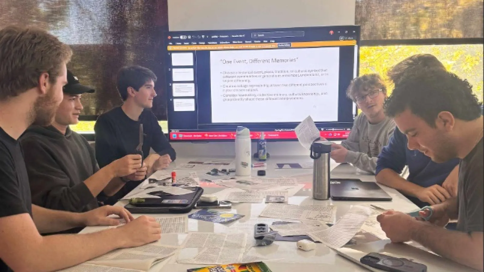 A group of students sitting around a table working on a collage, there is a screen in the background with information regarding the task.