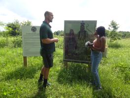 Badger Johnson and Angel Evans standing in front of Woodcock Nature Preserve's land acknowledgement as well as a woodcut portrait made by Suzanne Michele Chouteau
