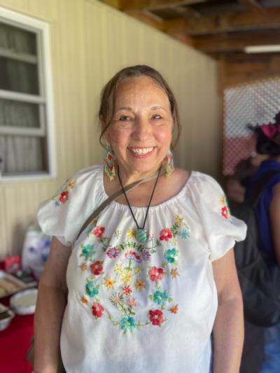 A portrait of Nancy Rose wearing a white shirt with short sleeves and colorful flower-embroidery lining the neck of the shirt. These colors match Nancy’s beaded earrings and necklace 