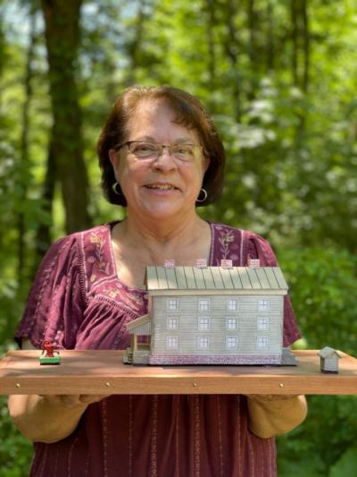 A portrait of Elaine Hutchinson holding a small diorama of the old boarding house, which sits on a thick wooden board. Elaine is wearing a maroon shirt with short sleeves that has white embroidery lining the neck of the shirt. Elaine has short brown hair and medium sized gold hoop earrings. The portrait is taken from the waist up.
