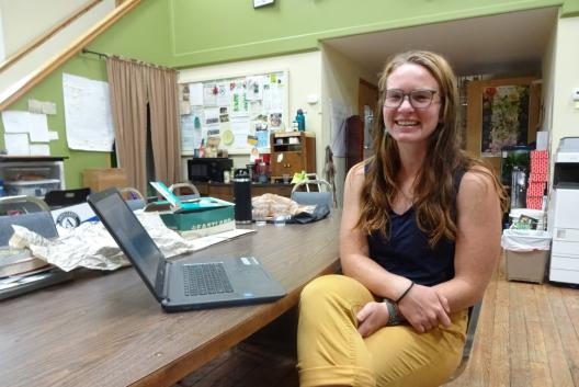 Madison Donohue sitting at a table with her laptop, smiling and facing the camera. She has long brownish reddish hair, and wears glasses, a blue sleeveless shirt, and yellow pants rolled up above her ankles. The office around Madison is covered with papers and office furniture.