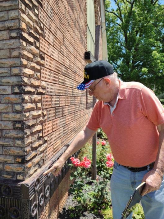 Larry Horn wearing a Veterans baseball hat, a salmon-colored t-shirt, and light blue jeans holding a clipboard and pointing towards a specific design on the Haydenville Methodist Church. Larry is pointing to the dark brown terracotta brick which is at waist height on the building. The rest of the building is red brick.