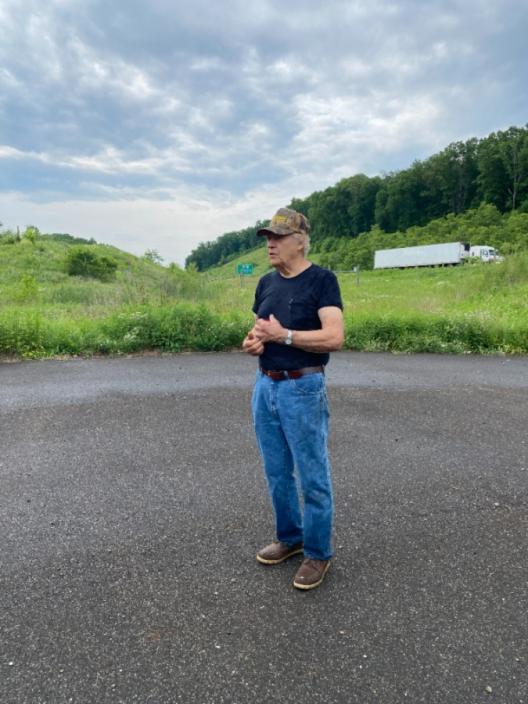 Pete Crane standing on the old Route 33, an abandoned highway, where his childhood home used to be. Pete is facing away from the camera and talking to someone out of frame. A large, white semi-truck is passing in the background, showing the new Route 33 and how close it is to the old one.