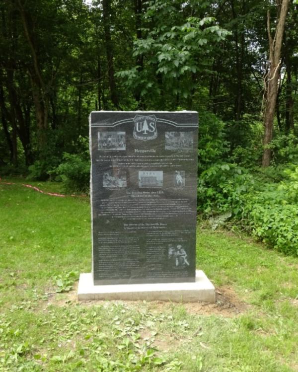 A tall, black stone monument with various photos and text engraved on its face. The monument sits on a white cement base, and it is surrounded by forest. The area around the monument is well-maintained grass.