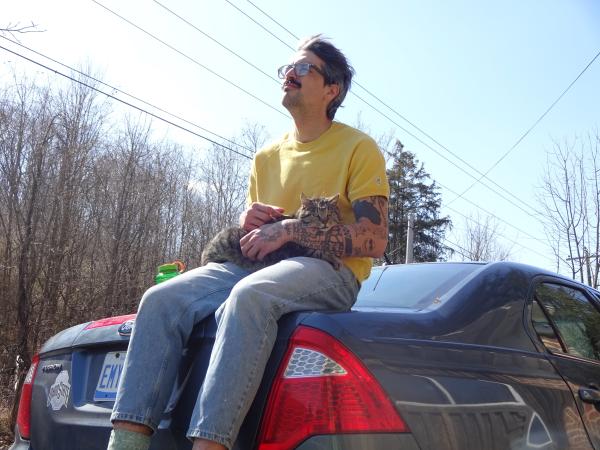 Andrew Mehall sitting on the back of his car and staring off into the distance with Chris Chmiel’s cat, Gus, on his lap.