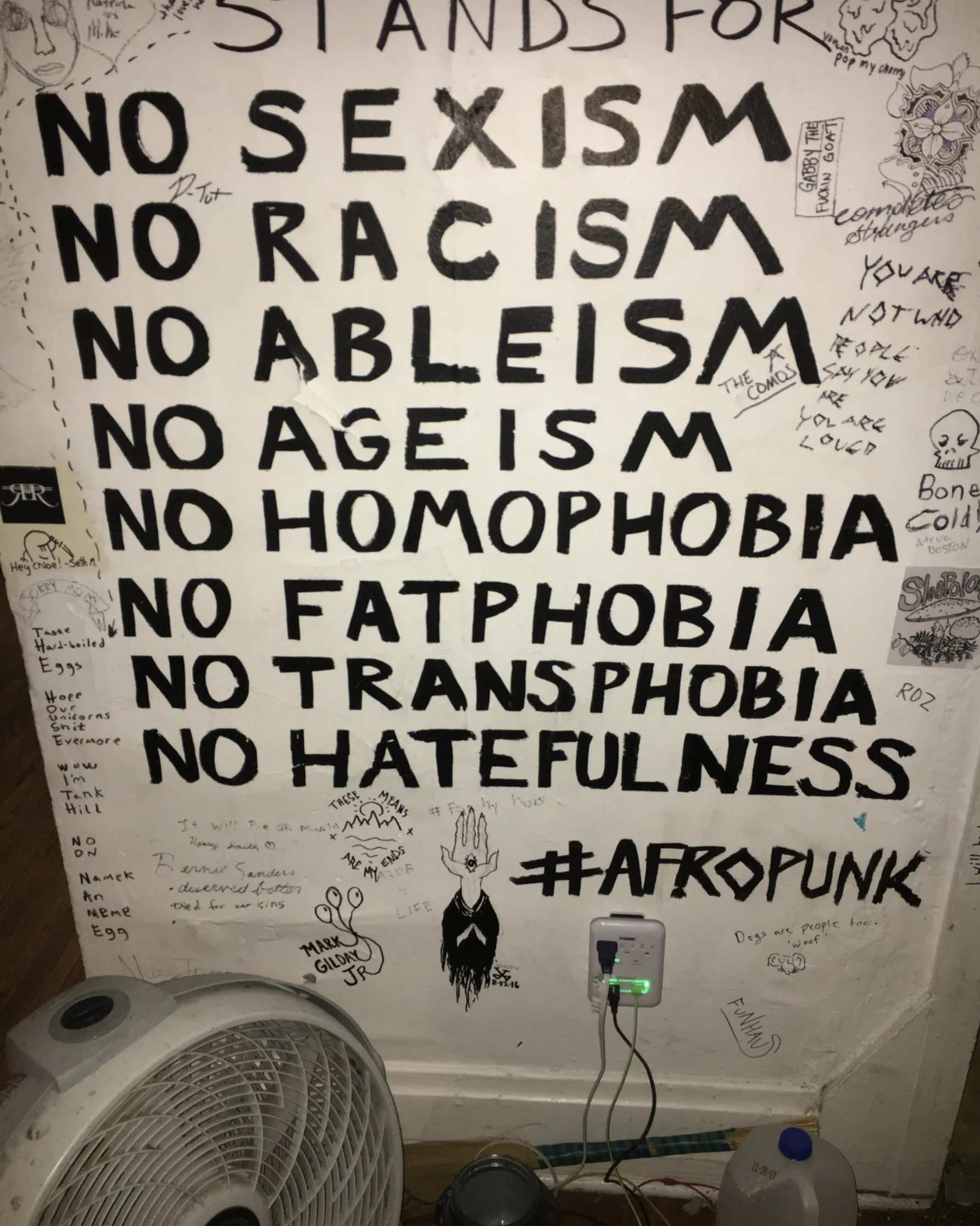 A wall with decorations and writing that says, "Stands for NO SEXISM, NO RACISM, NO ABLEISM, NO AGEISM, NO HOMOPHOBIA, NO FATPHOBIA, NO TRANSPHOBIA, NO HATEFULNESS #AFROPUNK."