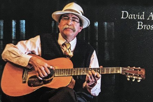 David Brose, a white man with a mustache and glasses wearing a white hat, sitting with a guitar in his lap