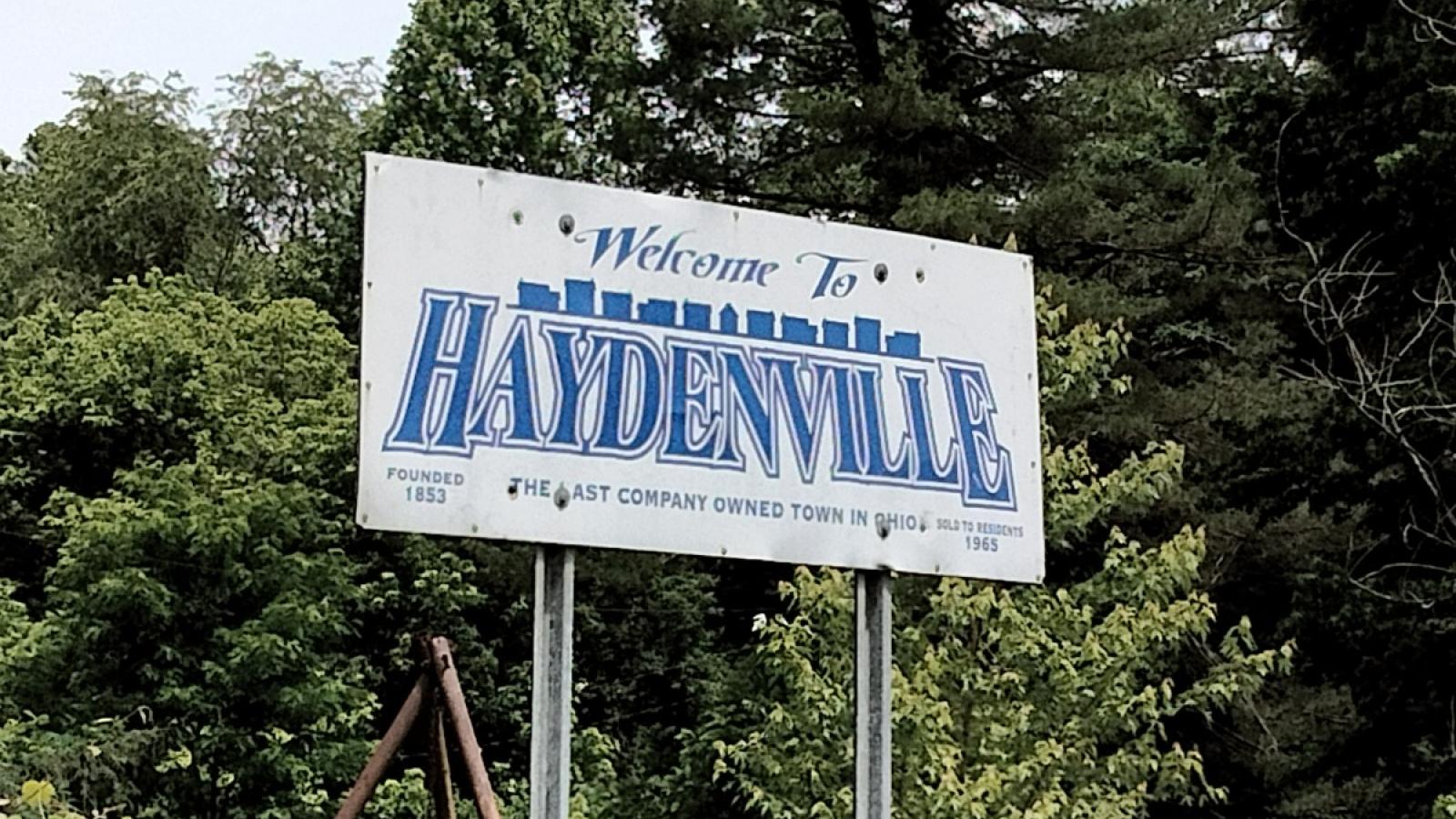 A sign saying "Welcome to Haydenville: the last company owned town in Ohio." The lettering is in blue against a white background, and the founding (1853) and selling (1965) dates of the town are located at the bottom of the sign. The sign stands high up, against a group of trees.