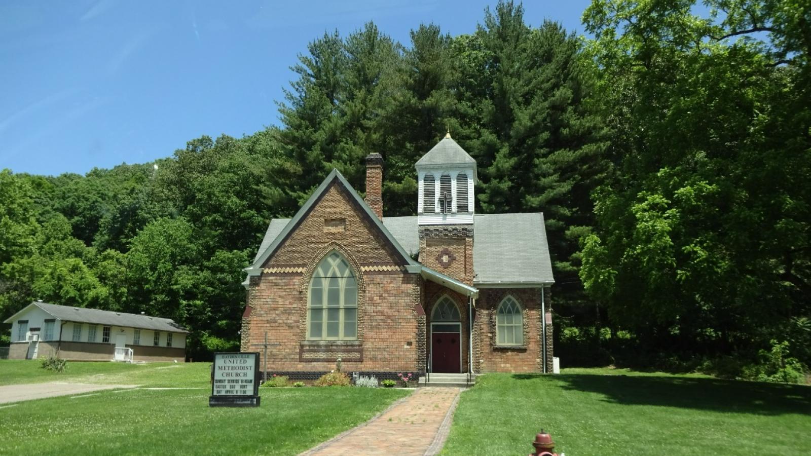 A small, brick Methodist church. The church has a large front window, a pair of red doors to the right of the large window, a chimney, and a white steeple with a cross on the front of it. There is a brick path leading up to the church building; on either side of the path is a well-manicured and very green lawn. On the left of the brick path is a white, square church sign which reads “Haydenville United Methodist Church, Sunday service 8:45AM, Easter egg hunt, Sat April 9. 1:00”