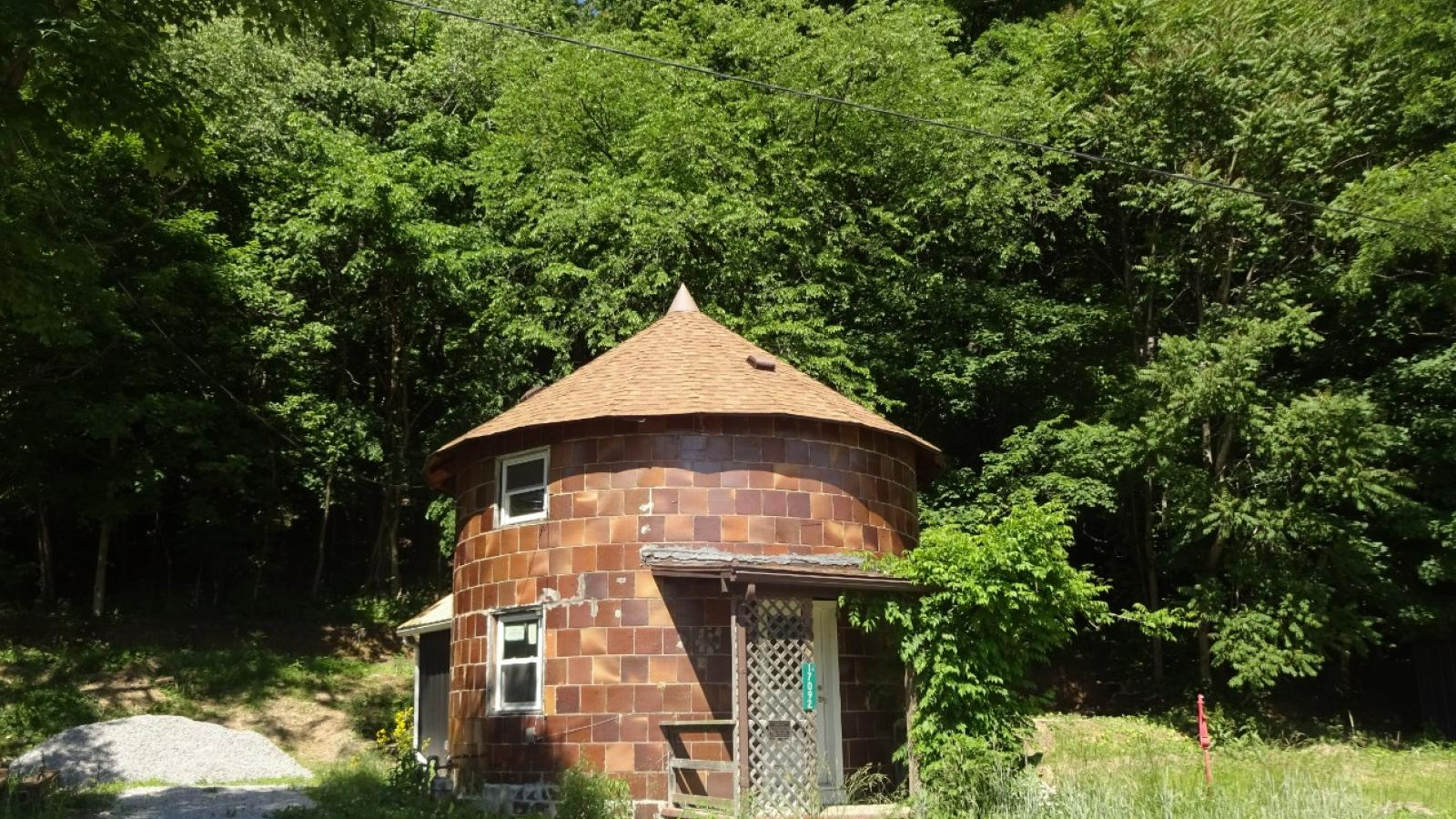 A round, silo-looking brick house with a pointed roof and curved, glazed, shiny brick walls. It is currently unoccupied and shows signs of neglect (e.g., no driveway or clear lawn). The round house is surrounded by the forest on all sides.