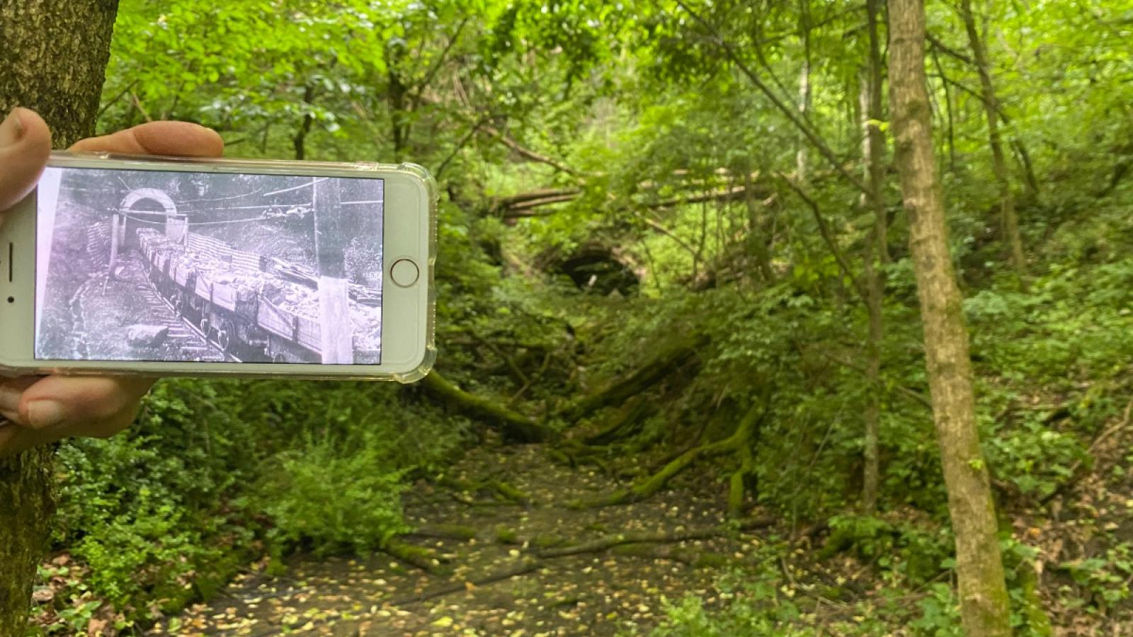 The hand of Pete Crane holding his iPhone which is displaying a black and white photo of the Haydenville tunnel when it was operating, carts coming in/out of its well-defined tracks. To the right of this photo is the old Haydenville tunnel today, barely visible behind the forest fauna and partially collapsed at the mouth. The train tracks are no longer visible, but it is apparent where the trail used to be.