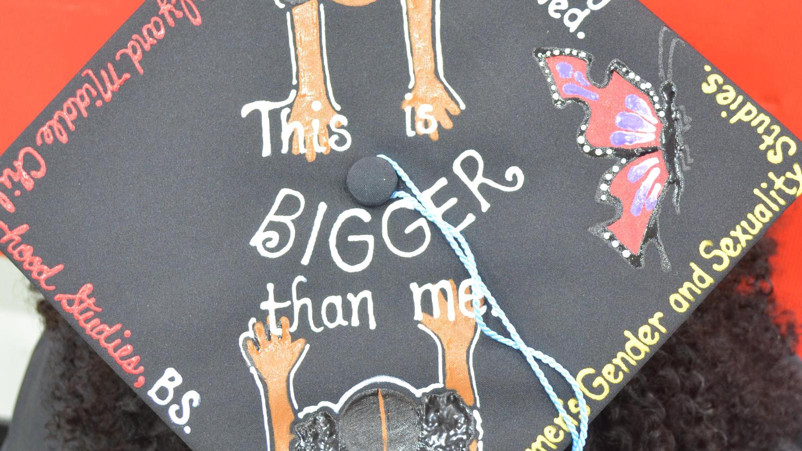 A graduating student wearing a mortarboard cap. In the center, it says "This is BIGGER than me." There are two girls reaching out toward each other from two corners of the bortarboard. The two girls are wearing shirts that say, "Black girl magic" and "I matter." Around the edges of the mortarboar, there is a butterfly and text that reads, "Beautifully flawed," "Women's gender and sexuality studies," "Early and middle childhood studies," "B.S.," and "Kines."