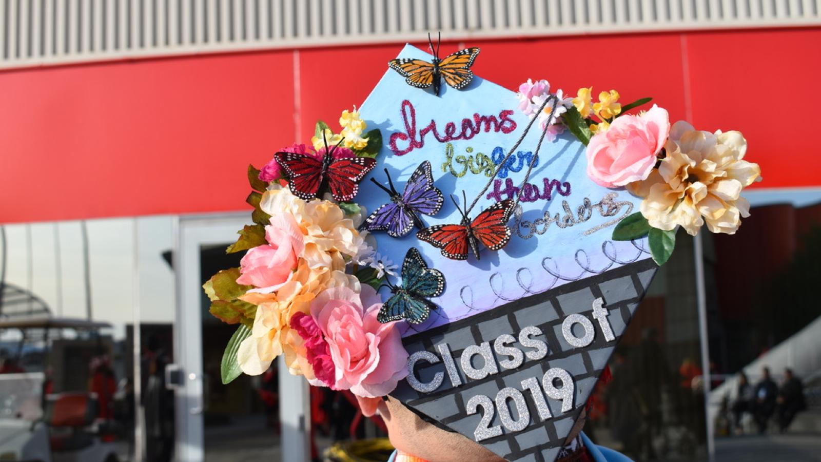 A graduating student wearing a mortarboard cap decorated with flowers and butterflies that says, "dreams biger than borders; Class of 2019"