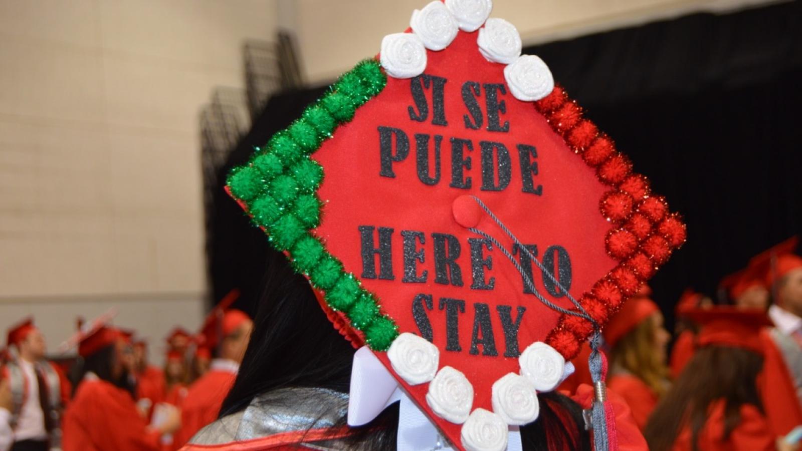 A graduating student wearing a green, white, red, and black mortarboard cap that says, "Si se puede; Here to stay"