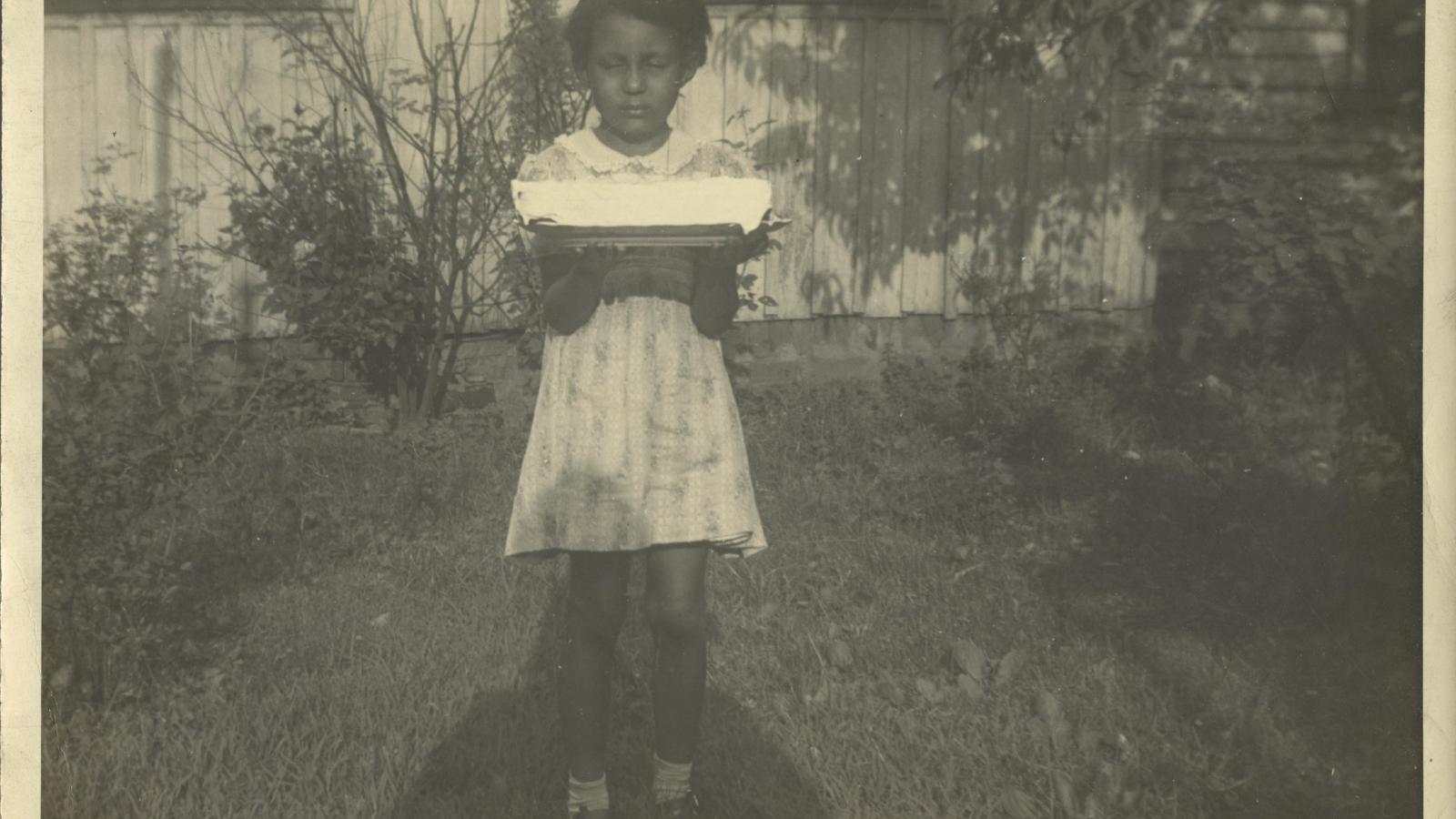 Photo of a young Geraldine Lewis holding a cake while standing in the backyard of 709 3rd Street