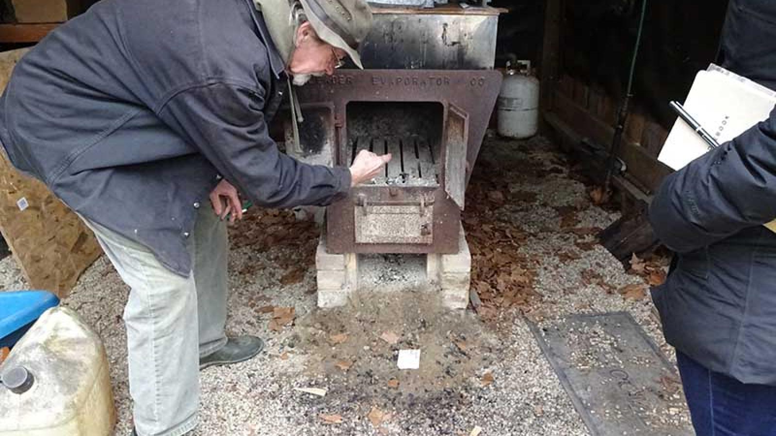 Ronny Richards leaning down while standing to the right and opening the door of cooking stove used in maple syrup production