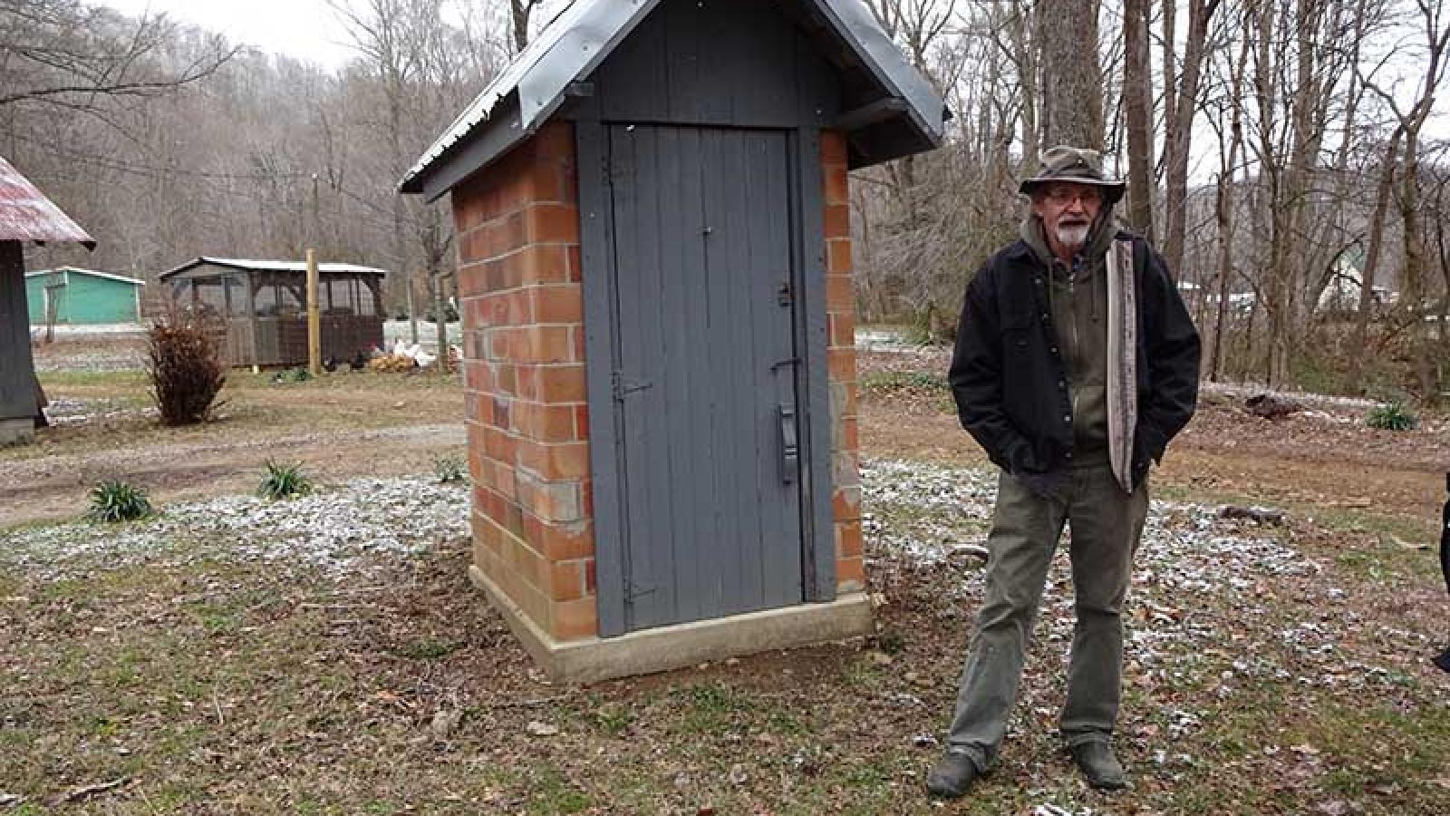 Ronny stands in front of a small red brick smokehouse with a grey roof and grey wooden door. 